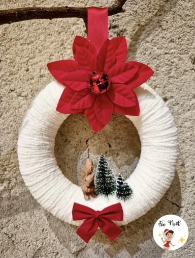 Couronne de Noël blanche avec une fleur rouge, sapins et une petite figurine.
