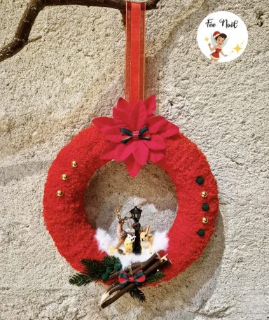 Couronne de Noël rouge décorée de fleurs et figurines sur un fond neutre.