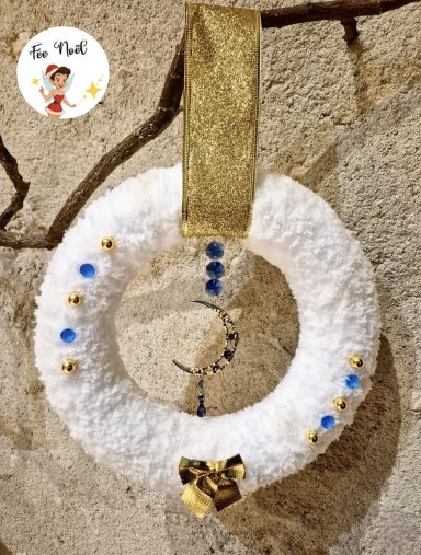 Couronne décorative blanche avec des perles bleues et un nœud doré suspendue au mur.